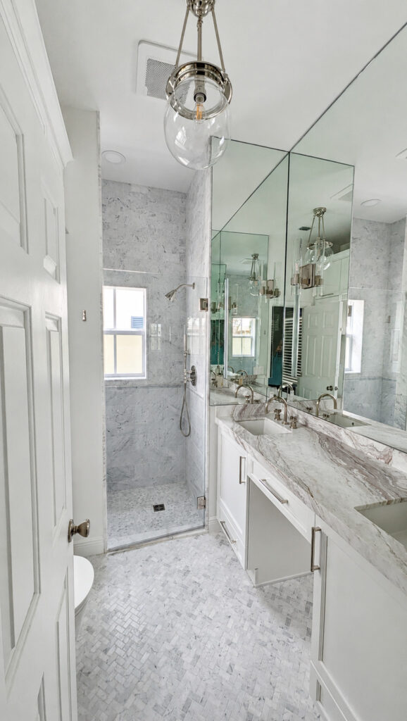 Renovated Boca Raton primary bathroom with a double quartzite-topped vanity, marble walk-in shower, and marble herringbone floor.