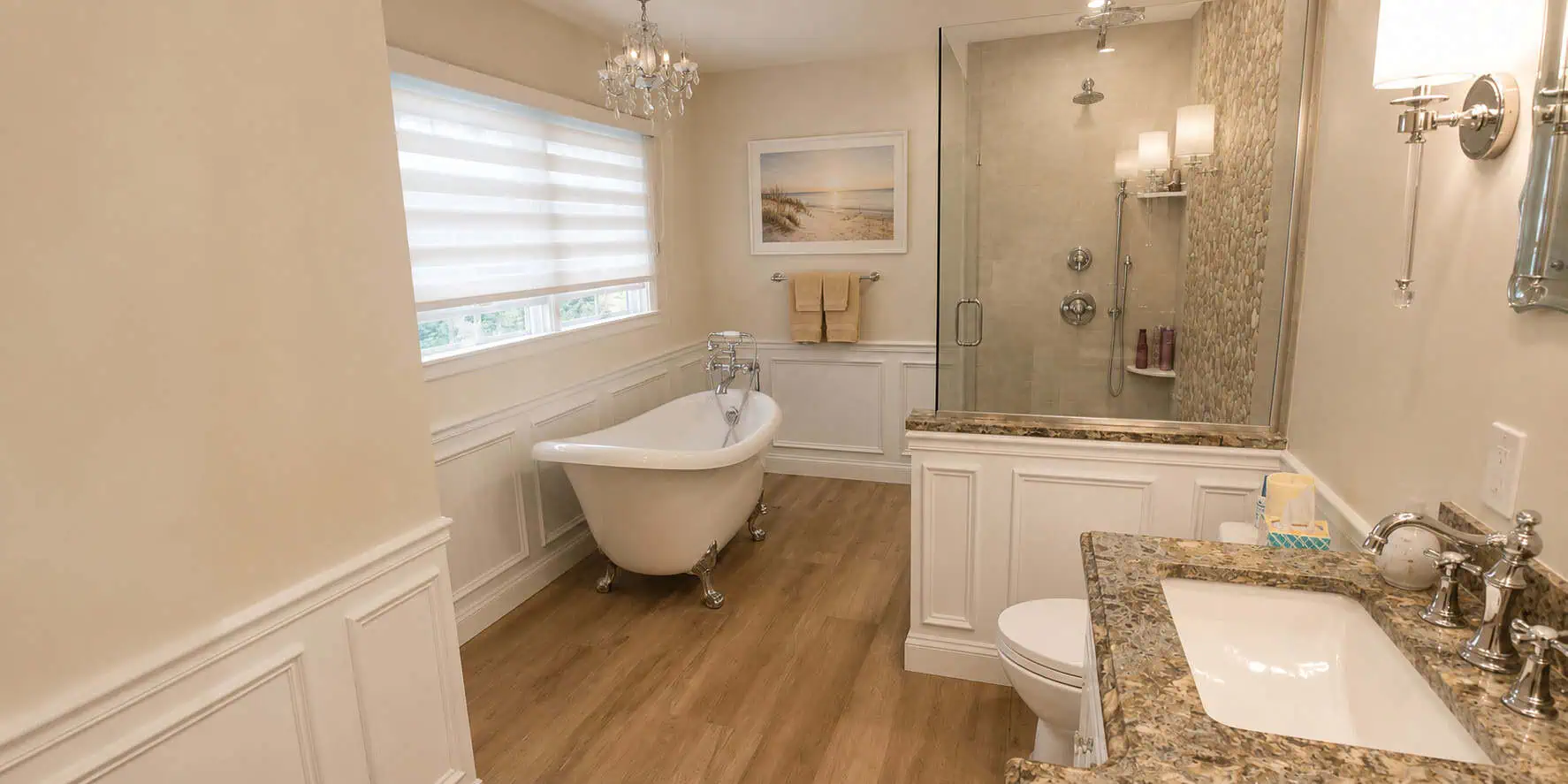 Primary bathroom remodel with a slipper clawfoot tub beneath a crystal chandelier, a glass walk-in shower with a stone mosaic wall, and raised-panel wainscoting.