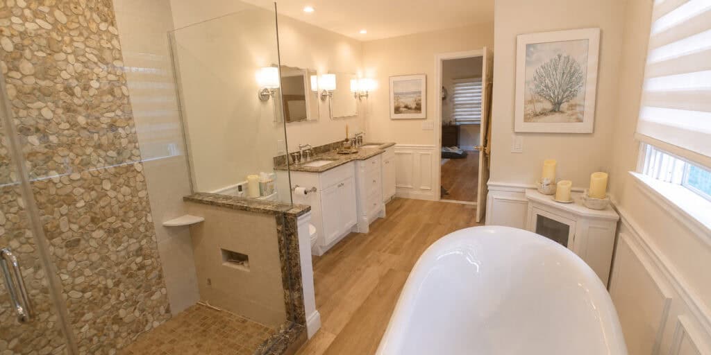 Primary bathroom remodel featuring a full-height river-stone mosaic shower wall, a granite double vanity, a slipper clawfoot tub, and raised-panel wainscoting.