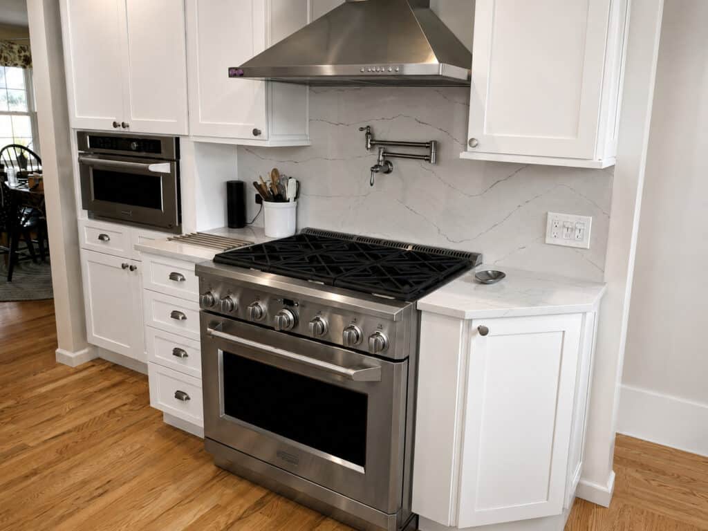 Compact white shaker kitchen with a stainless pro-style gas range, chimney hood, pot filler, and a veined slab backsplash.