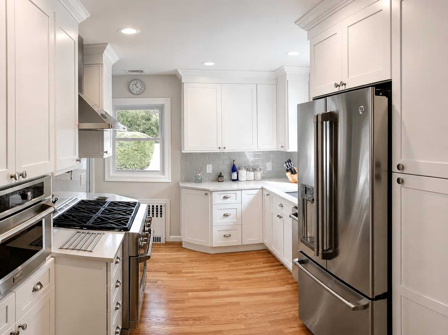 Wide view of a compact white shaker galley kitchen with stainless appliances, a grey subway tile backsplash, and hardwood floors.
