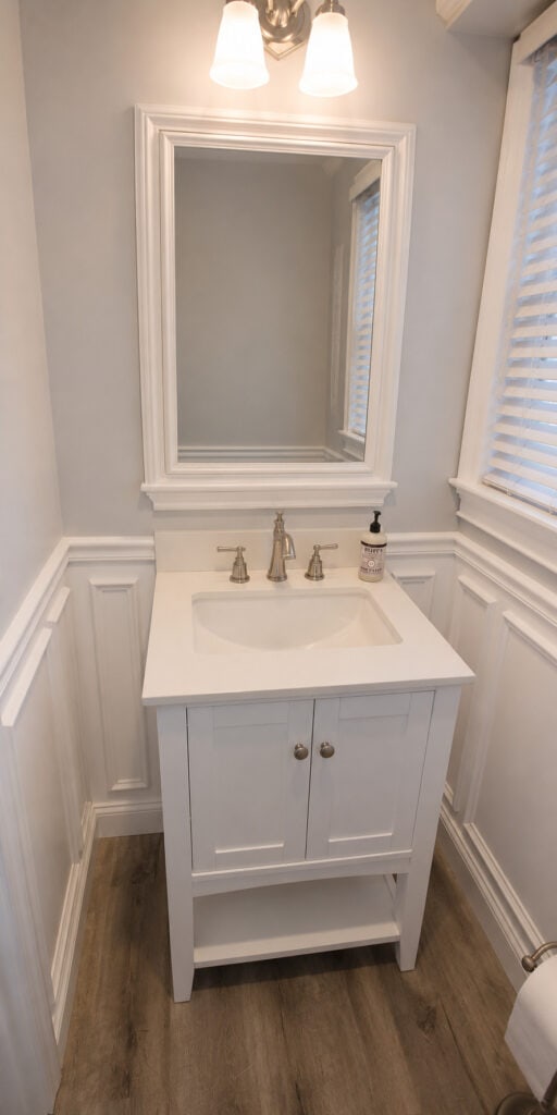 Compact powder room vanity with a white cabinet, white countertop, brushed nickel faucet, a framed mirror, and raised-panel wainscoting.