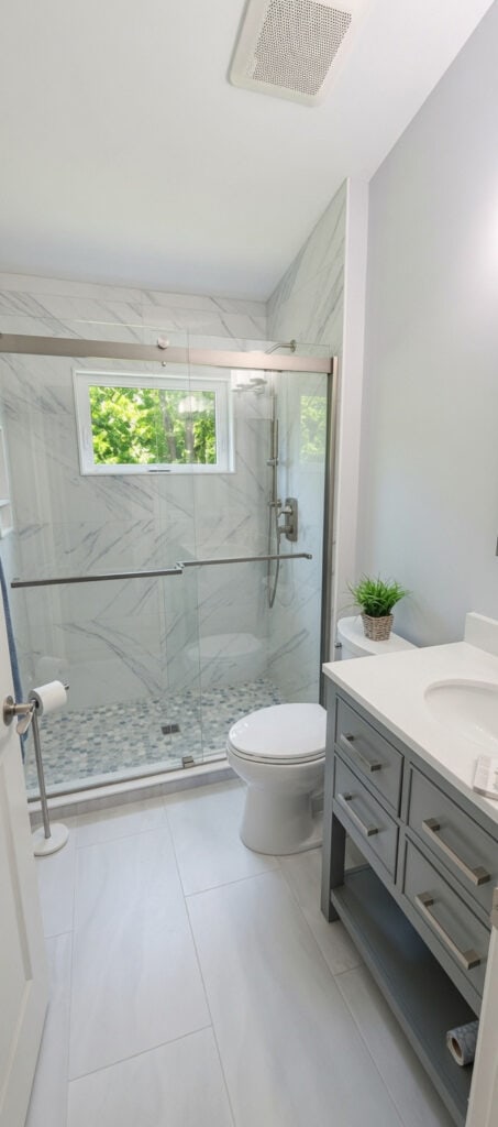 Lake Worth bathroom addition with a marble-look porcelain tile shower, a glass sliding door, an impact awning window, and a grey vanity with a quartz top.