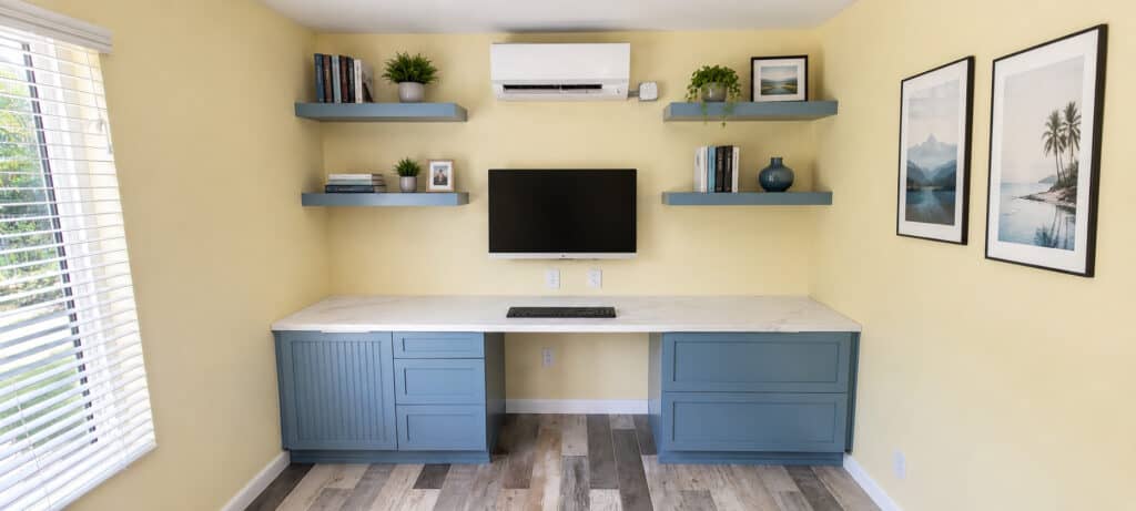 Coastal home office with a built-in blue desk, matching floating shelves, and a wall-mounted monitor set around a ductless mini-split.
