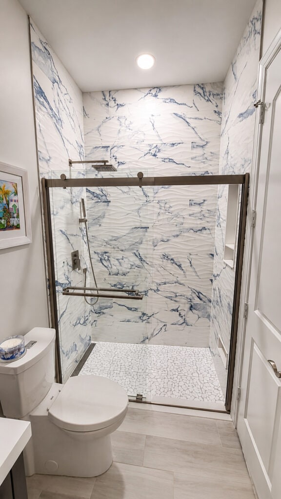 Ocean Ridge coastal bathroom with a blue-and-white marble-look shower, a wavy dimensional tile accent, a pebble mosaic floor, and a sliding glass door.