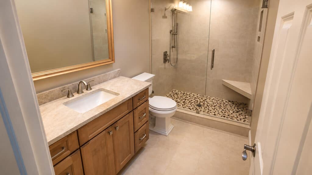 PGA National bathroom with a wood vanity, quartz top, gold-framed mirror, and a pebble-floor walk-in shower with a corner bench.
