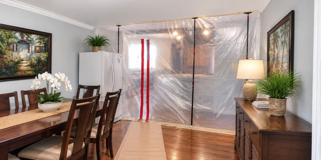 Zippered plastic dust barrier sealing off a kitchen from a dining room, with kraft-paper floor protection, during a home remodel.
