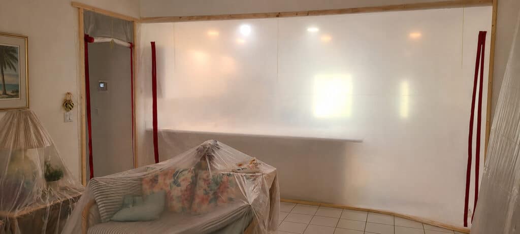 Furniture covered in plastic and a poly containment wall with zippered access during a home remodel.