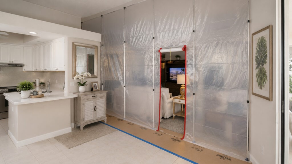 A pole-mounted plastic dust barrier isolating a living room from a kitchen work area, with floor protection, during a remodel.