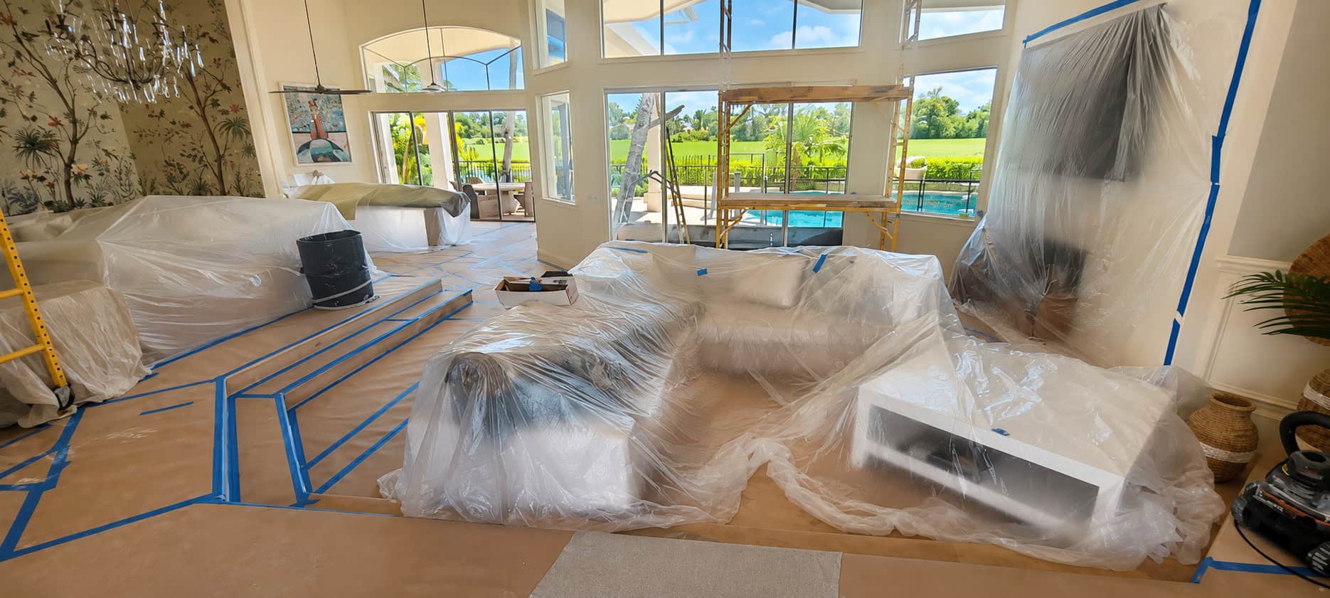 A luxury living room with furniture wrapped in plastic and kraft-paper floor protection during a whole-home remodel.