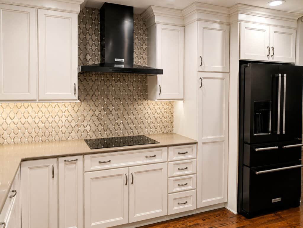 Timeless transitional kitchen range wall with an arabesque mosaic backsplash, a black chimney hood, glazed cream cabinets, and a black stainless refrigerator.