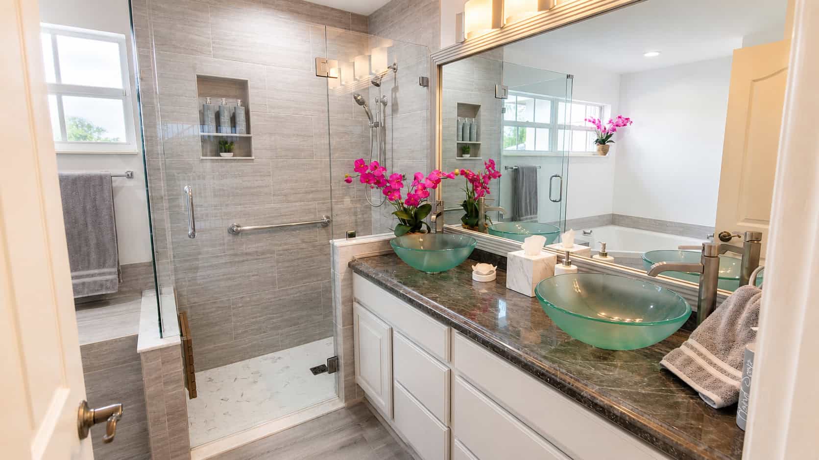 Wellington primary bathroom with a frameless glass walk-in shower, grey wood-look tile, a double vanity with glass vessel sinks, and a soaking tub.