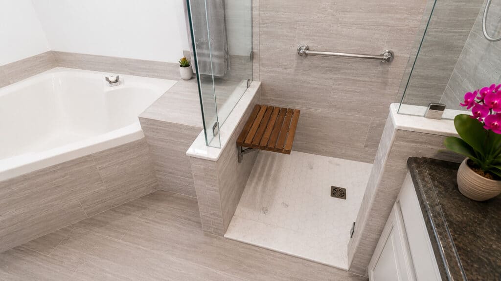 Wellington bathroom shower with a teak fold-down seat, a hex marble mosaic floor, grab bars, and grey wood-look tile beside a soaking tub.