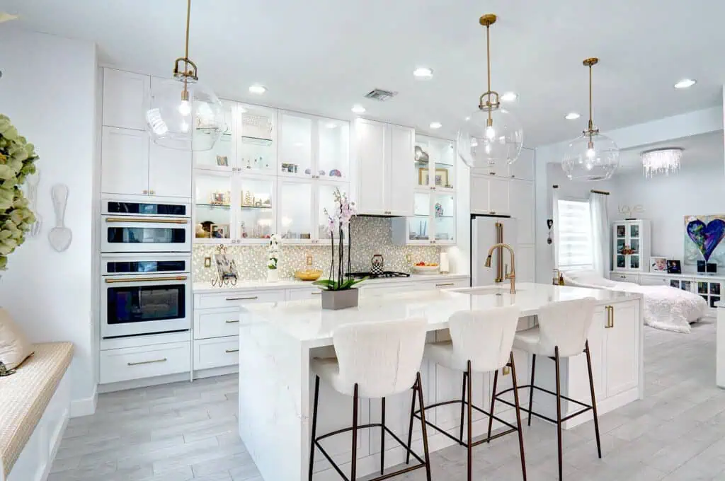 Palm Beach County kitchen with a white quartz waterfall island, glass-front lit cabinets, white double ovens with brass handles, and glass globe pendants.