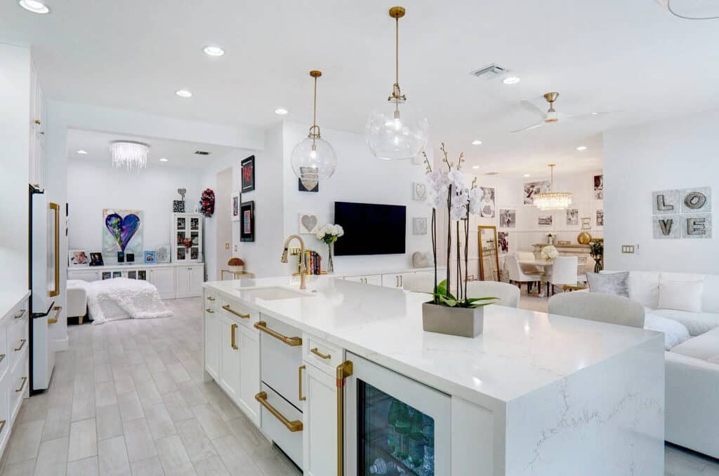 Open-concept Palm Beach County kitchen with a white quartz waterfall island, brass hardware, and glass globe pendants, open to a living area.
