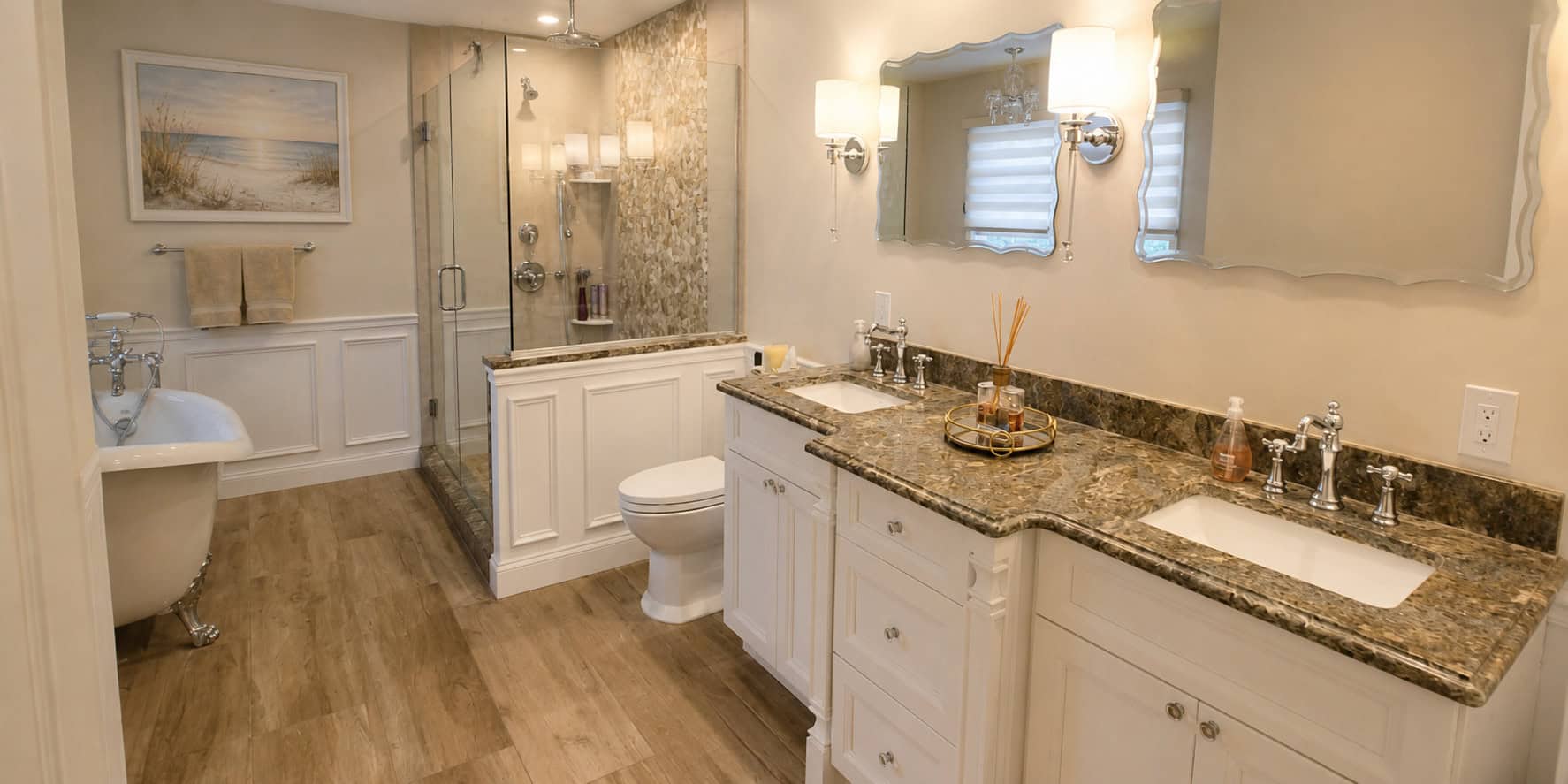 Spa-style primary bathroom remodel with a slipper clawfoot tub, a glass walk-in shower with a river-stone mosaic feature wall, and a granite double vanity framed by raised-panel wainscoting.