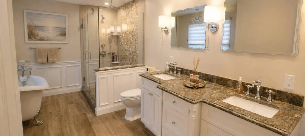 Primary bathroom remodel with a granite double vanity under a framed mirror and wall sconces, a glass walk-in shower with a stone mosaic wall, and a slipper clawfoot tub beyond.