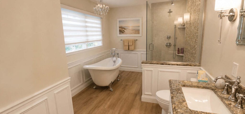 Primary bathroom remodel with a slipper clawfoot tub beneath a shuttered window, a glass walk-in shower with a stone mosaic feature wall, and a granite vanity, framed by raised-panel wainscoting.