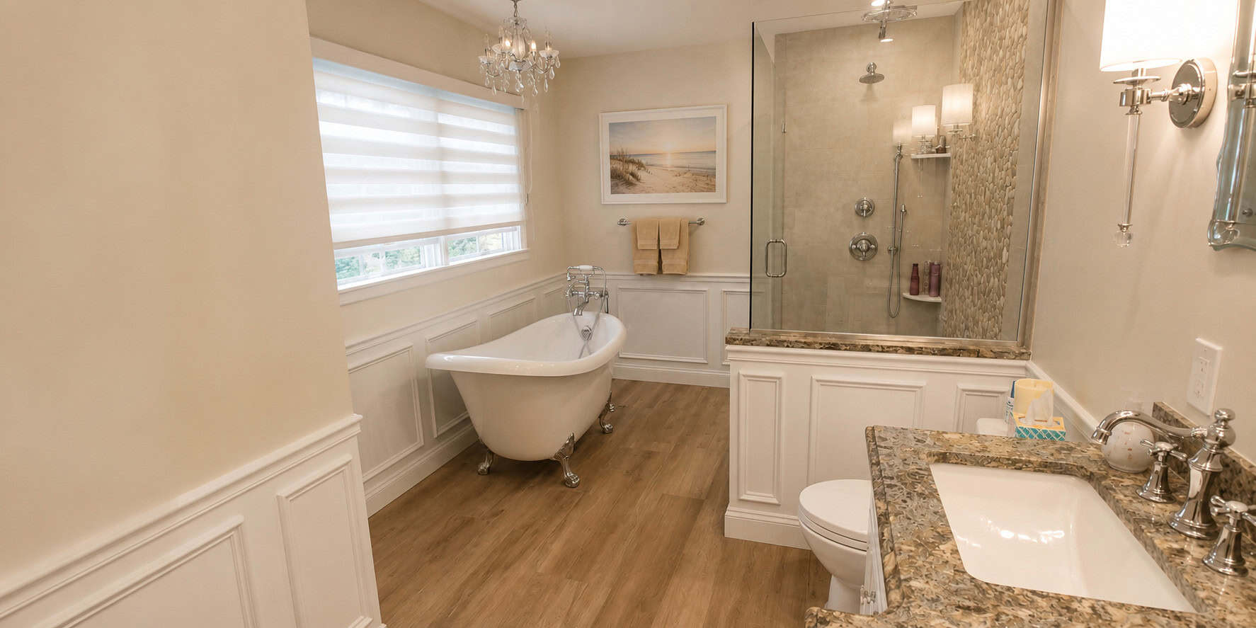 Primary bathroom remodel with a slipper clawfoot tub beneath a crystal chandelier, a glass walk-in shower with a stone mosaic wall, and raised-panel wainscoting.