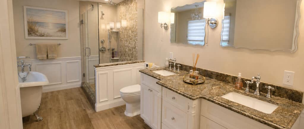 Spa-style primary bathroom remodel with a slipper clawfoot tub, a glass walk-in shower with a river-stone mosaic feature wall, and a granite double vanity framed by raised-panel wainscoting