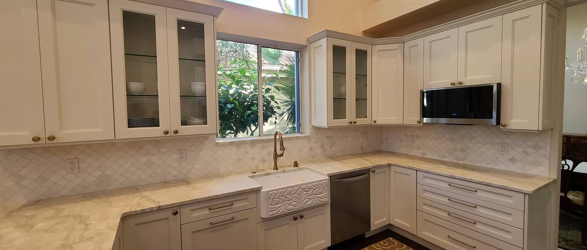 BallenIsles kitchen remodel in Palm Beach Gardens with cream cabinetry, brass hardware, quartzite countertops, an arabesque tile backsplash, a carved apron-front farmhouse sink and glass-front display cabinets
