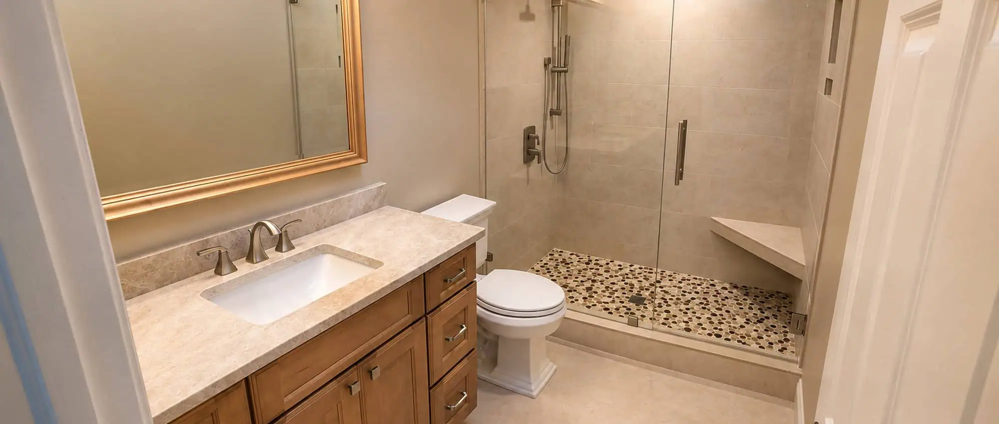 Palm Beach Gardens PGA bathroom remodel with a wood-toned vanity, a framed mirror and a frameless glass shower with a pebble-mosaic floor and a corner bench