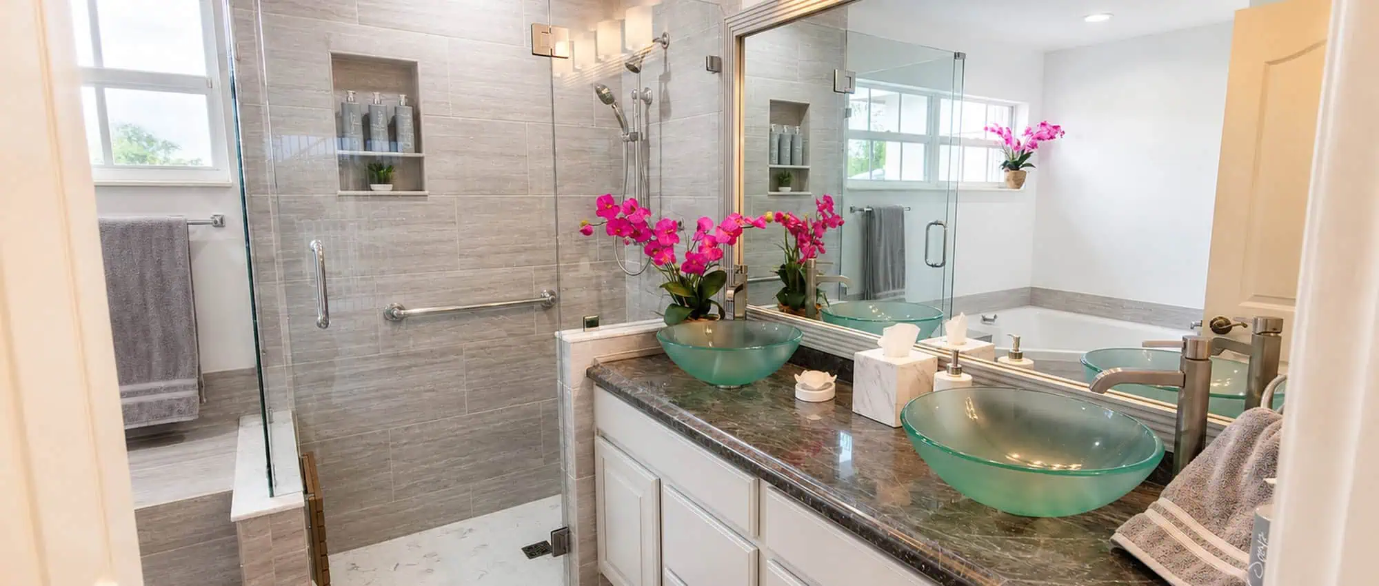 Wellington primary bathroom remodel with a glass walk-in shower, a tiled shower niche, twin green glass vessel sinks on a dark stone-look vanity and a large framed mirror