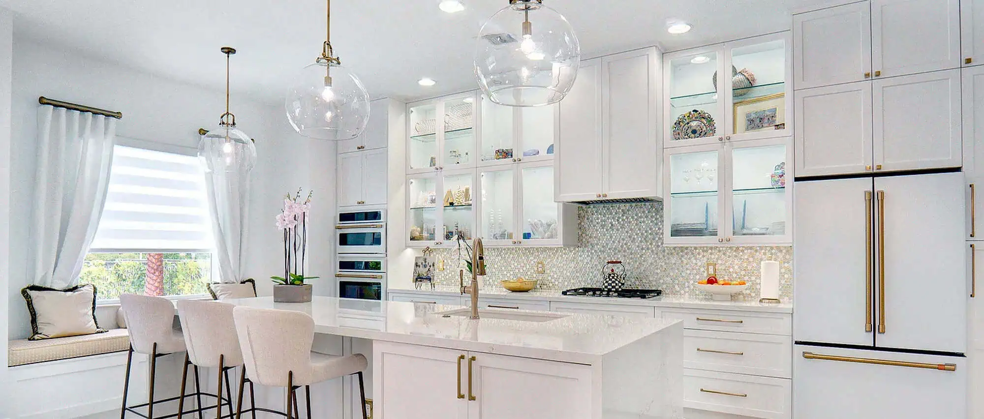 Palm Beach County kitchen remodel with white cabinetry, brass hardware, a pearl mosaic backsplash, glass-front lit upper cabinets, a waterfall-edge island and a built-in window seat